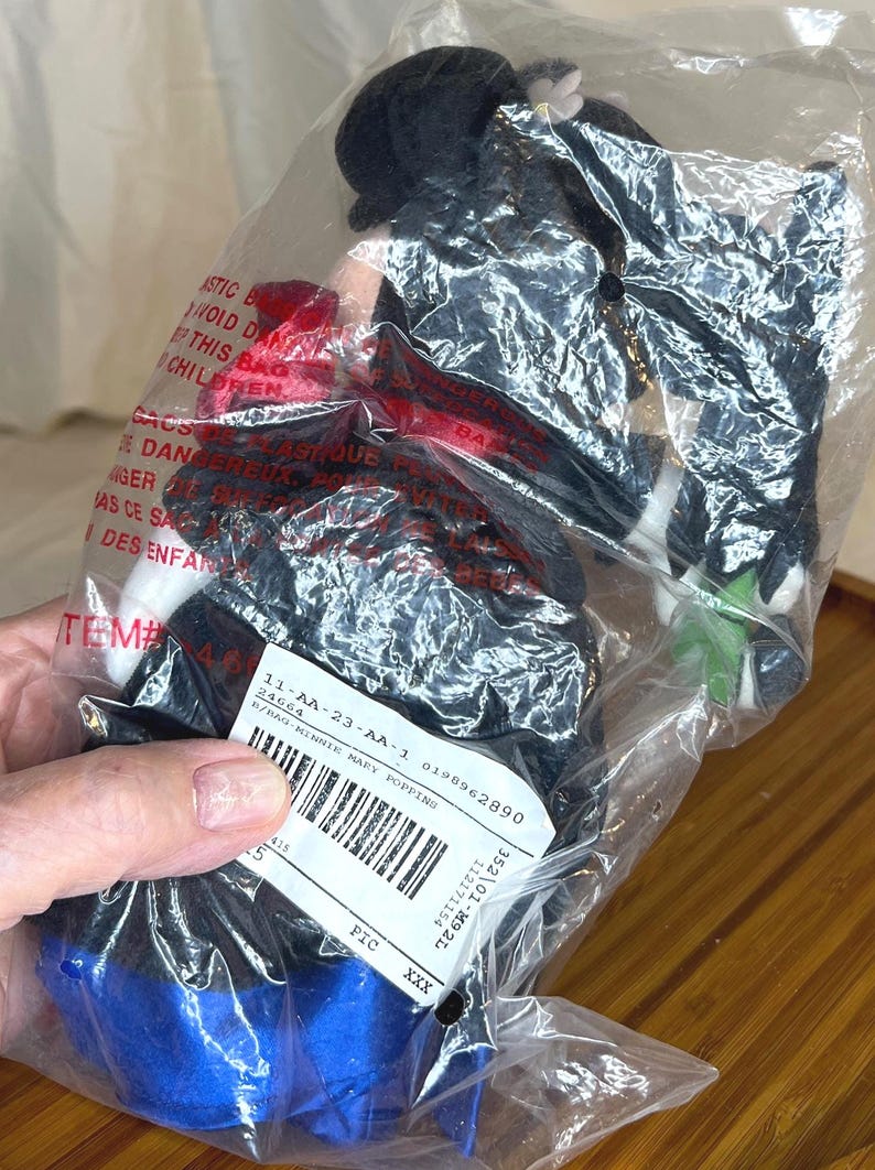 K&ouml;nnte beinhalten: Eine Minnie Mouse Pl&uuml;schpuppe in einer Plastikt&uuml;te. Die Puppe tr&auml;gt einen schwarzen Hut, ein schwarzes Oberteil und einen blauen Rock. Ein Etikett mit Barcode und Text ist an der T&uuml;te befestigt. Die Puppe ist teilweise sichtbar.