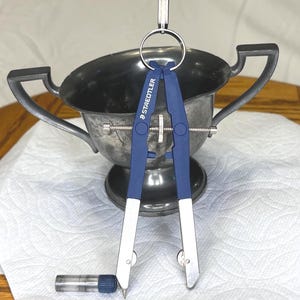Puede incluir: Un compás Staedtler azul y plateado con un recipiente para minas de lápiz. El compás está suspendido frente a un cuenco de metal plateado con asas. El compás tiene un tornillo plateado y un anillo en la parte superior para colgar.