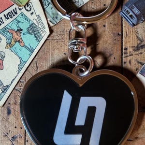 May include: Heart-shaped keyring with a black background and a white stylized "LN" logo. The keyring has a silver-coloured metal ring and clasp. The background includes vintage paper ephemera on a wooden surface.