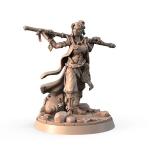 May include: A tan-colored miniature figurine of a warrior holding a staff. The figure is wearing a cloak, armor, and pants. The base of the figurine has a sculpted face and rocks. The warrior has a determined expression.