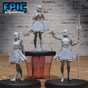 May include: Three miniature figurines of a woman wearing a maid outfit. The woman on the left is walking, the woman in the middle is standing, and the woman on the right is holding a staff. All three figurines are painted in a light gray color.