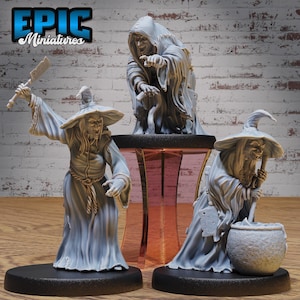 May include: Three painted miniature figures of witches wearing pointed hats and robes. The witches are posed in different positions, one holding a cauldron, one holding a knife, and one holding a staff. The figures are painted in shades of gray and white.