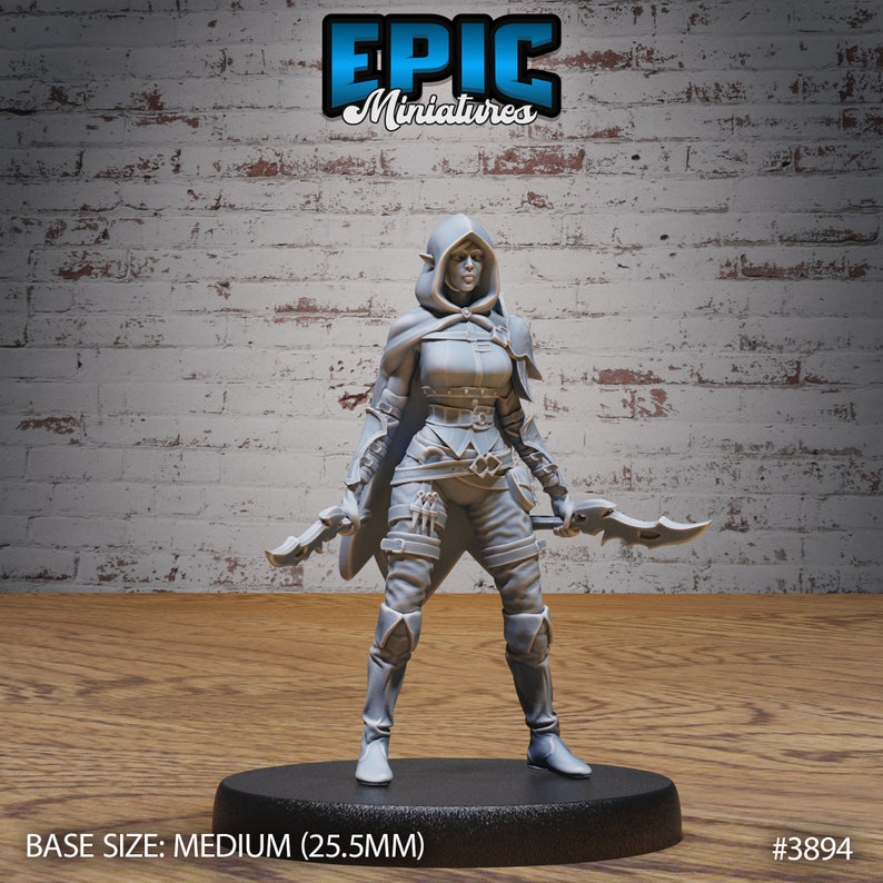 Puede incluir: Figurilla miniatura de pl&aacute;stico gris de una elfa femenina con una capa con capucha y sosteniendo dos dagas. La miniatura est&aacute; de pie sobre una base redonda con el texto "BASE SIZE: MEDIUM (25.5MM)" y "#3894" impreso en ella.