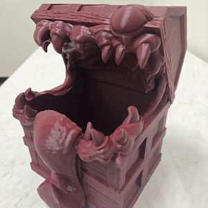 Puede incluir: Un cofre del tesoro burdeos impreso en 3D con un diseño monstruoso. La tapa y los lados del cofre están detallados con texturas de tablones de madera. La parte superior presenta una boca monstruosa con dientes afilados.