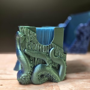 May include: A teal and blue card holder featuring an octopus design. The holder has a textured, wood-like appearance with tentacles wrapping around the sides. The interior holds a stack of blue cards. A second holder is visible in the background.