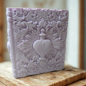 May include: A lavender-colored, ornate book-shaped object with intricate floral and heart-shaped designs. The object has a detailed, embossed surface and appears to be made of a solid material. The book is resting on a wooden surface.