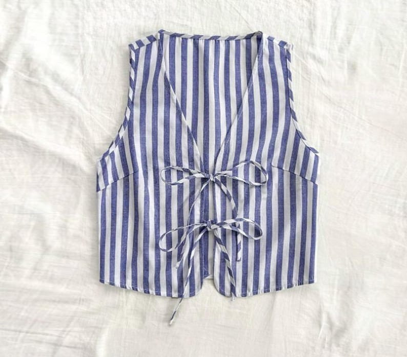 May include: A sleeveless, blue and white striped waistcoat with a V-neck and a tie closure at the front.