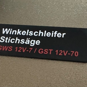 Könnte beinhalten: Schwarzes Etikett mit weißem Text, der "Winkelschleifer Stichsäge GWS 12V-7 / GST 12V-70" lautet.