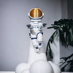 Può includere: Una statuetta di astronauta bianca con un casco dorato è in piedi su una base a forma di razzo bianca con nuvole bianche. L'astronauta indossa una tuta spaziale blu e bianca e ha le braccia distese.