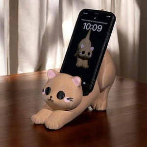 May include: A tan cat-shaped phone holder with pink ear and cheek details. A phone, showing a cat image, is in the holder. The phone displays the time as 10:09. The holder is on a wooden surface.