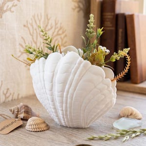May include: A white seashell-shaped vase filled with greenery and small white flowers. The vase sits on a wooden surface with seashells, a tag, and a gold ring. The background includes books and a curtain with a coral pattern.