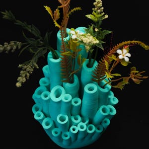 Peut inclure: Un vase turquoise en forme de corail avec diverses ouvertures, rempli de verdure artificielle et de fleurs blanches. Le vase a une surface texturée et est placé sur un fond noir, créant un contraste saisissant. L'arrangement évoque un thème naturel sous-marin.