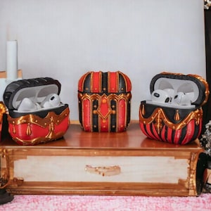 May include: Three red and gold treasure chest-shaped cases for wireless earbuds. The cases have black and gold trim and are sitting on a wooden shelf.