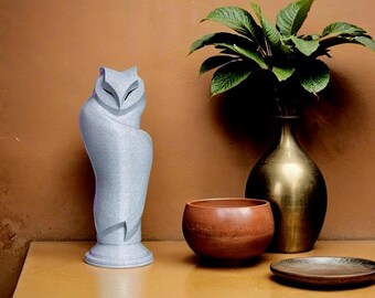 May include: A silver owl sculpture with a smooth, textured surface. The owl is perched on a wooden table with a brown bowl and a gold vase with green leaves.