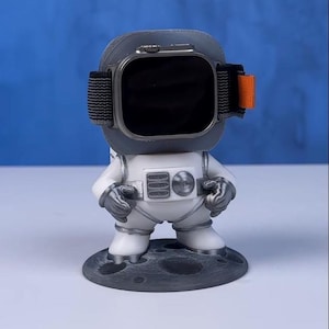Könnte beinhalten: Eine weiße Astronautenfigur mit einer grauen Mondbasis, die eine schwarze Smartwatch mit einem orangefarbenen Band hält.