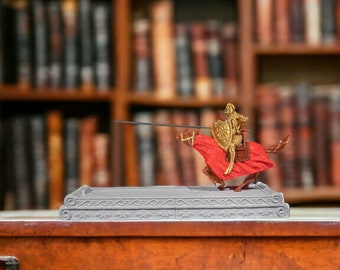 Puede incluir: Una figurita de caballero dorado y rojo montando un caballo, sosteniendo una lanza y un escudo, sobre una base de piedra gris. La figurita está delante de una estantería con muchos libros.
