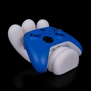 以下が含まれることがあります： 白のスタンドに置かれた青と白のゲームコントローラー。 コントローラーは黒いボタンと白い底面を備えています。 スタンドはコントローラーを保持するように設計されています。