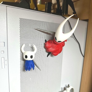May include: Two character figurines, one white with a red cloak and the other white with a blue cloak, are attached to a computer. The figurines have a sword. The figurines are from the video game Hollow Knight.