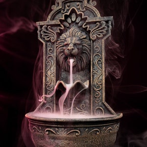 Op de afbeelding: Een decoratieve wierookbrander met een leeuwenkoptuit, omgeven door rook. De brander is gemaakt van donker, verweerd metaal met ingewikkelde details. De rook is wit en wolkig en creëert een mystieke sfeer.