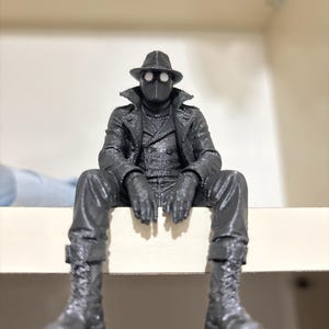 May include: A dark gray 3D printed figurine of a character wearing a fedora, a long trench coat, and lace-up boots. The figure has a mask with two prominent white circular eye lenses and is posed in a seated position with legs dangling downward.