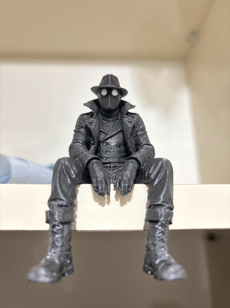 May include: A dark gray 3D printed figurine of a character wearing a fedora, a long trench coat, and lace-up boots. The figure has a mask with two prominent white circular eye lenses and is posed in a seated position with legs dangling downward.