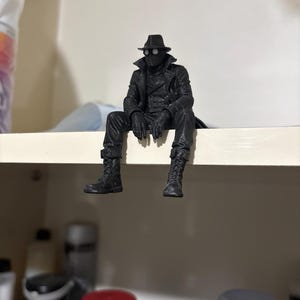 May include: A black figurine of a character wearing a fedora, a long trench coat, gloves, and lace-up boots. The figure is depicted in a seated position with legs dangling, featuring white circular lenses over the eyes and a textured matte finish.