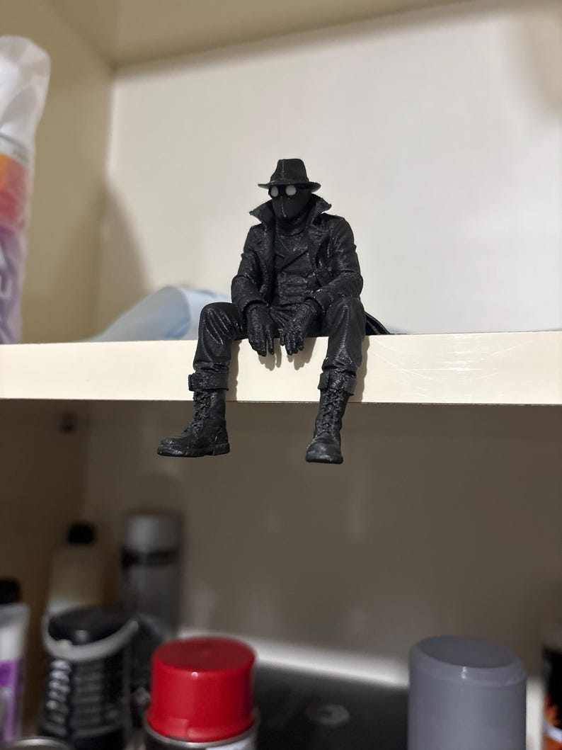 May include: A black figurine of a character wearing a fedora, a long trench coat, gloves, and lace-up boots. The figure is depicted in a seated position with legs dangling, featuring white circular lenses over the eyes and a textured matte finish.