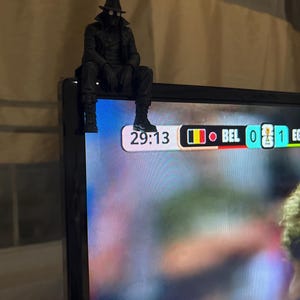 May include: A black figurine of a character wearing a fedora, trench coat, and goggles, perched on the top corner of a television screen. The screen displays a sports scoreboard showing 29:13, the country code BEL, and a score of 0 to 1.