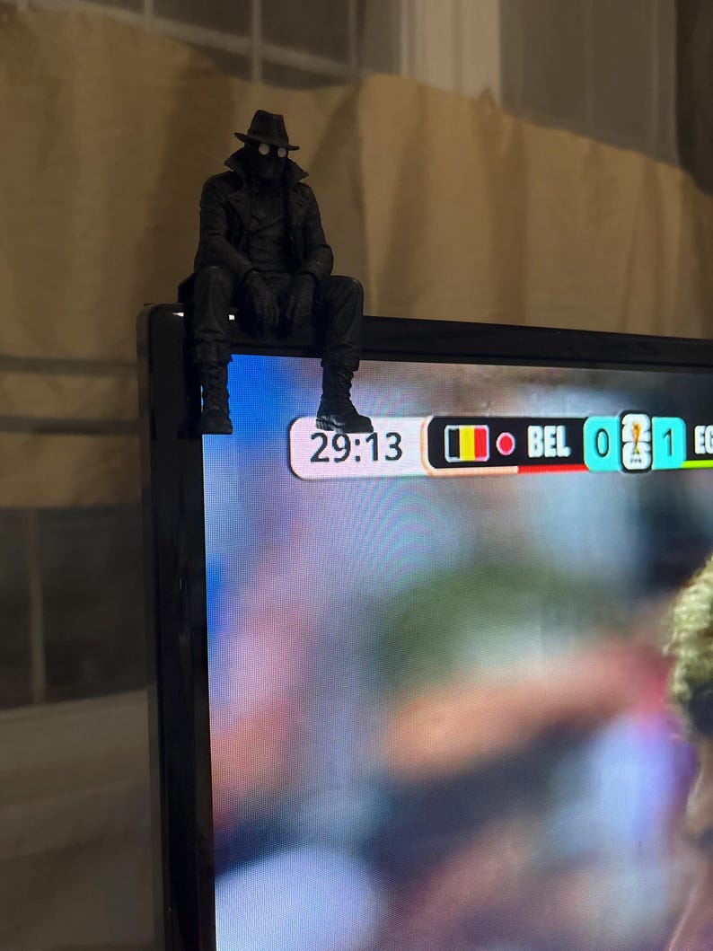 May include: A black figurine of a character wearing a fedora, trench coat, and goggles, perched on the top corner of a television screen. The screen displays a sports scoreboard showing 29:13, the country code BEL, and a score of 0 to 1.