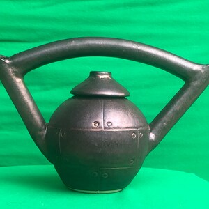 May include: A dark gray ceramic teapot with a unique design. The teapot has a round body, a lid, and a large, arched handle. The surface has a textured appearance with small, rivet-like details. The spout is angled, and the base is a lighter color.