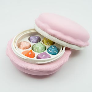 May include: A pink dice case shaped like a macaron, with a white interior and a lid. Inside are seven colorful dice in various colors, including orange, green, and blue. The dice are numbered with white numerals.