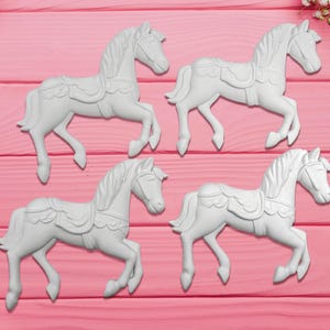 May include: Four white carousel horse figurines arranged on a pink wooden surface. Each horse is sculpted with detailed manes, saddles, and bridles. The horses are in a trotting pose, suggesting movement and a classic design.