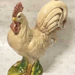 May include: A ceramic rooster figurine featuring a cream-colored body with a textured plumage, a prominent red comb and wattle, and a yellow beak. The bird stands on a green, leaf-shaped base with detailed clawed feet, showcasing a glossy finish.