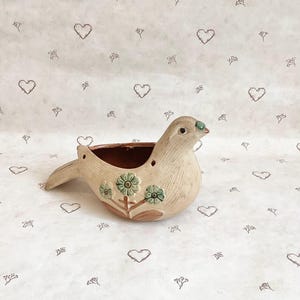 May include: A ceramic bird-shaped planter in beige with a brown interior. The bird has a green beak and green floral accents. The planter is on a white surface with heart and floral patterns.