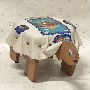 May include: A wooden footstool shaped like a turtle with a light-colored fabric cover featuring a colorful graphic print of a landscape with a rainbow and clouds. The stool has four wooden legs and a head with a single visible googly eye.
