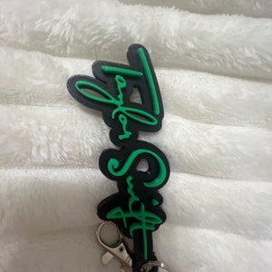 May include: Black and green keychain with the text "Taylor Swift" in a stylized font.