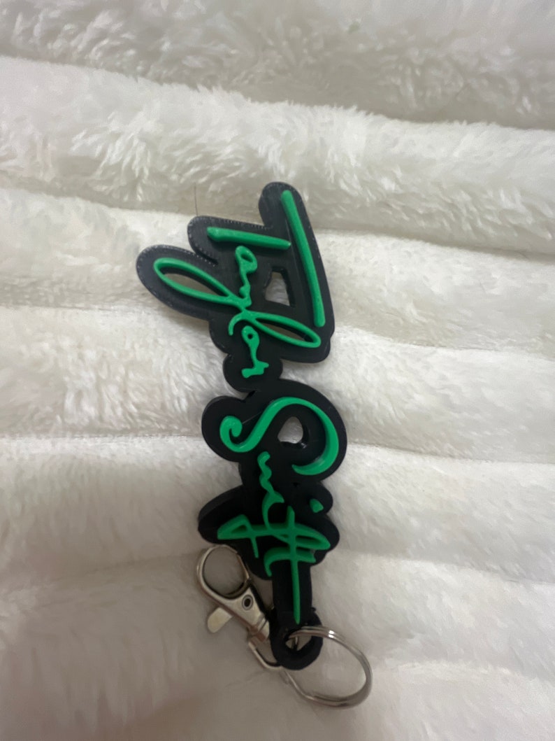 May include: Black and green keychain with the text "Taylor Swift" in a stylized font.