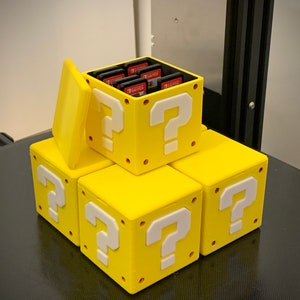 May include: A stack of yellow question mark blocks with a white question mark on each side. The top block has a lid that is open, revealing a compartment for storing Nintendo Switch game cartridges.