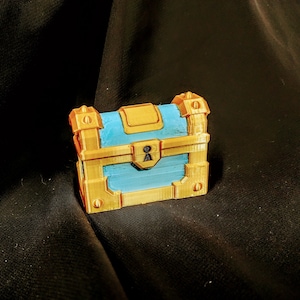 May include: A small, gold-colored treasure chest with a blue lid and a keyhole. The chest has decorative details and appears to be made of plastic.
