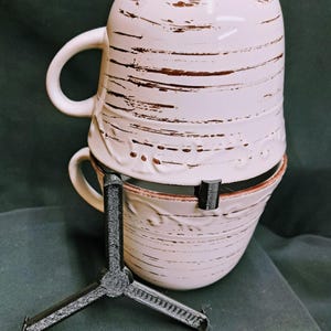 May include: Two stacked white ceramic mugs with brown distressed lines and a decorative rim. A black tripod-like stand supports the bottom mug. The mugs have a handle on the side. The background is a dark green fabric.
