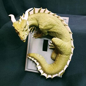 May include: A green dragon figurine with white spines, curled around a light switch plate. The dragon has a detailed textured body and a curled tail.