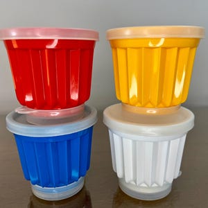 May include: Four colorful, stackable plastic containers with lids. The containers are red, yellow, blue, and white. Each container has a ridged design and a clear, round lid. The containers are sitting on a brown surface.