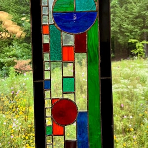 May include: A rectangular stained glass panel featuring a geometric composition of clear, green, blue, red, and black glass pieces. The design includes two prominent circular shapes, one blue and one red, integrated into a grid of vertical and horizontal lines.