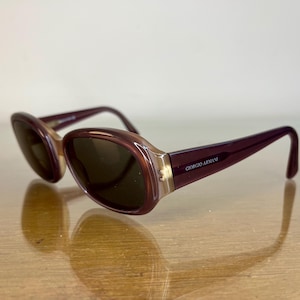 May include: Giorgio Armani sunglasses featuring oval-shaped frames in a deep burgundy and translucent beige gradient. The temples are a solid dark burgundy with the brand name Giorgio Armani printed in silver on the side. The lenses are dark brown.