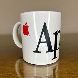 May include: A white ceramic mug featuring a red apple logo on the left side and the black capital letter A followed by a lowercase p on the right side. The mug has a curved handle on the left and a smooth, cylindrical body with a glossy finish.