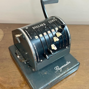 May include: An antique Paymaster cash register in a dark gray finish. The register features a handle, dollar and cent indicators, and the word "Paymaster" in cursive. The register is on a wooden surface.