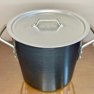 May include: A large, aluminum cooking pot with a lid. The pot is dark gray with two curved handles and a silver lid with a handle. The pot is designed for cooking and food preparation.