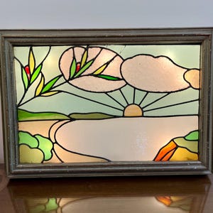 Pode incluir: Painel retangular de vitral retratando um pôr do sol sobre um corpo de água. A obra de arte apresenta uma moldura de madeira, um sol amarelo, nuvens brancas e folhagem verde e amarela. O vidro é iluminado por trás, criando um brilho quente.