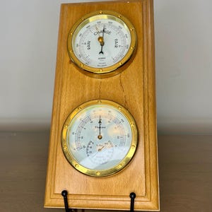 Puede incluir: Estación meteorológica de madera con dos medidores circulares. El medidor superior indica las condiciones meteorológicas "Change" y "Fair", mientras que el medidor inferior muestra la temperatura y la humedad. Los medidores tienen bordes dorados.