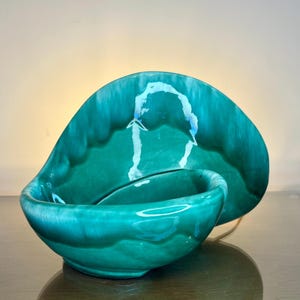 May include: A teal-colored ceramic bowl and decorative plate set. The bowl is a rounded shape with a smaller, attached bowl. The plate is a large, curved shape. The set has a glossy finish and a unique, organic design.